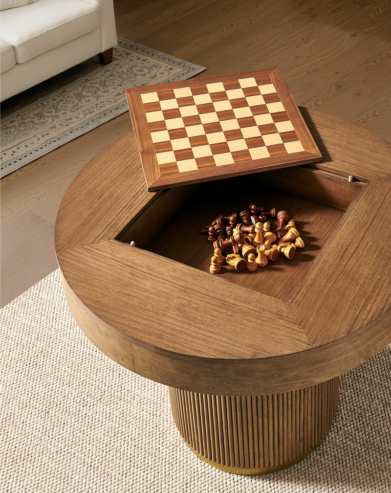 A round wooden coffee table with a lift-top chess game storage feature, shown in a warm living room setting with a sofa and rug in the background.