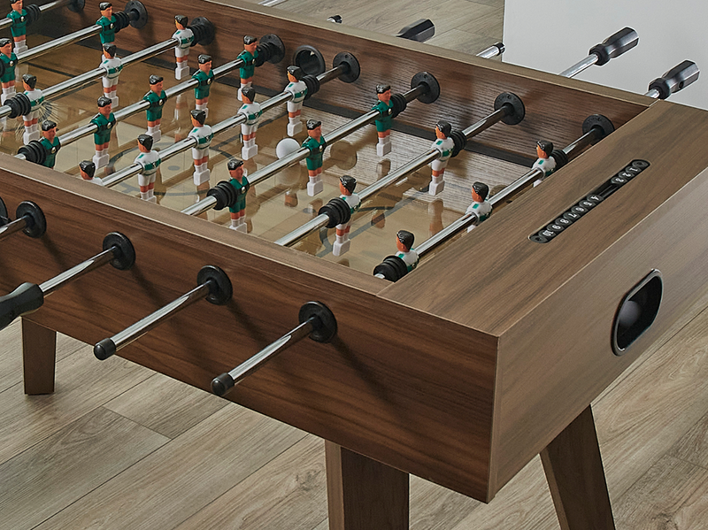 A wooden foosball table with silver metal rods, black handles, and small player figures, shown on a light wood floor.